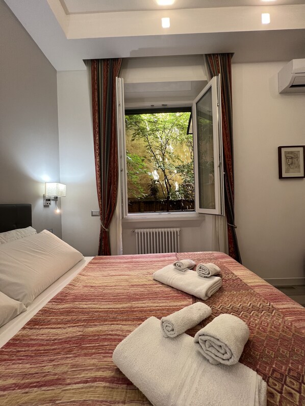 A hotel room with a bed, a window with curtains, a radiator, and a wall-mounted air conditioner.
