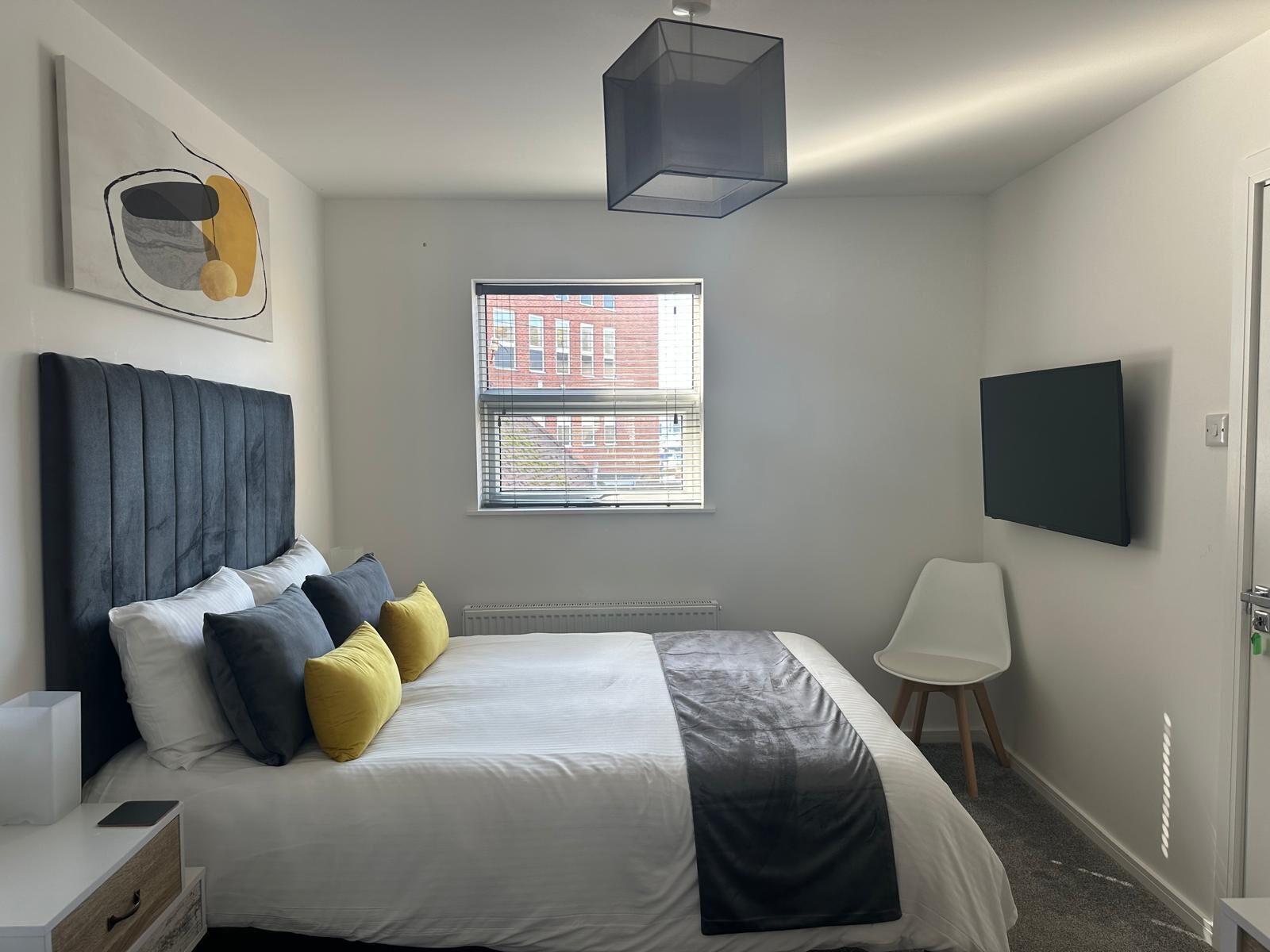 A bedroom with a bed, bedside table, lamp, window, and a TV mounted on the wall.