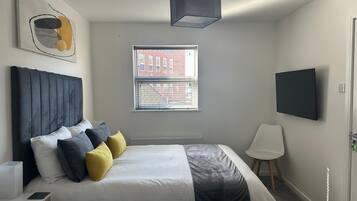 A bedroom with a bed, bedside table, lamp, window, and a TV mounted on the wall.