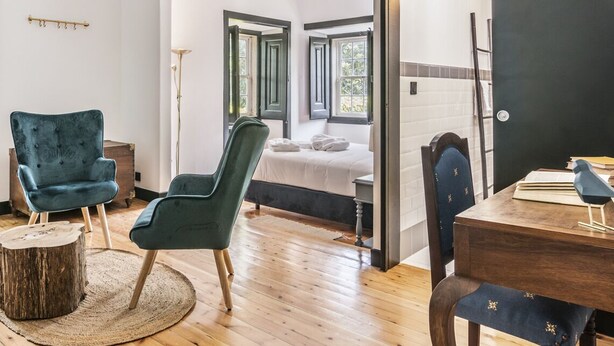 A modern hotel room with a wooden floor, two green armchairs, a round wooden table, a bed with white linens, and a desk with books and a lamp. - Quinta dos Lobos Boutique Hotel - Nature Experience (Sintra)