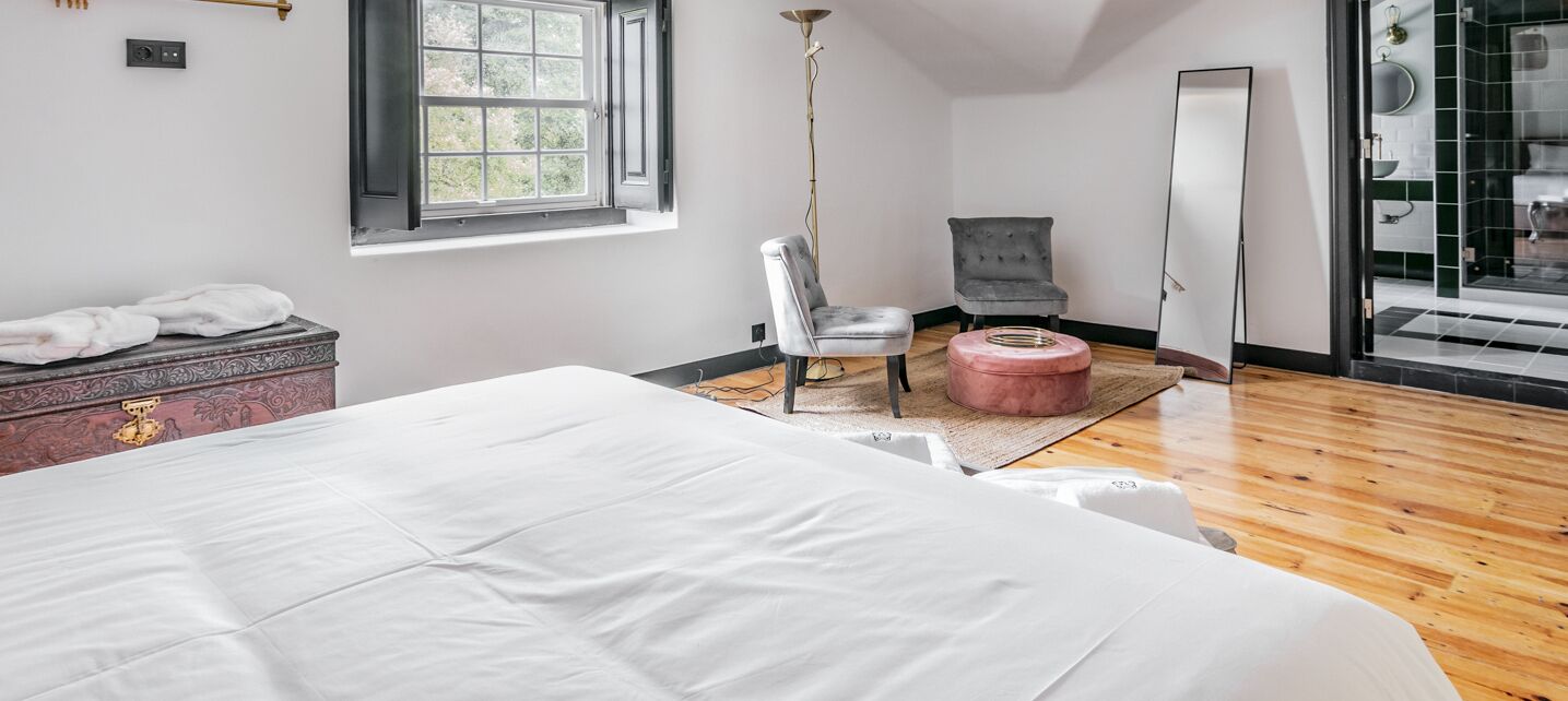 Quarto com uma cama grande, uma cadeira, um banquinho pequeno, cômoda e espelho.