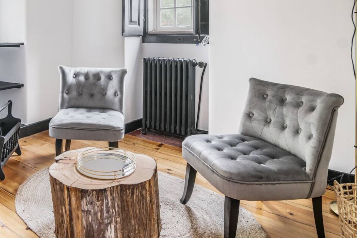 Um quarto com duas poltronas cinzas, uma mesa redonda de madeira, uma cadeira de metal preta e um banquinho azul. Há janelas com molduras pretas, um radiador e piso de madeira.