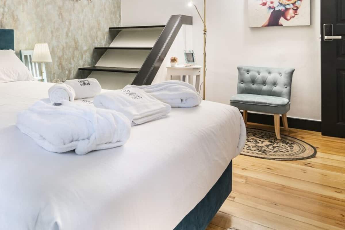 Cama bem arrumada com lençóis e toalhas brancas, cabeceira azul, uma cadeira e uma mesa pequena com abajur.