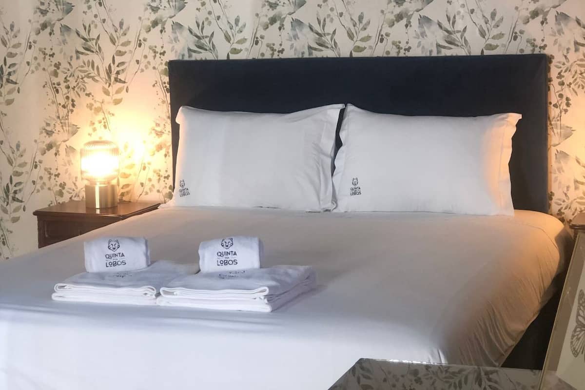 Uma cama bem arrumada com lençóis e almofadas brancas, uma mesinha de cabeceira com uma toalha dobrada e um objeto decorativo, e uma parede com padrão floral.