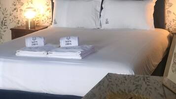 Una cama bien hecha con sábanas y almohadones blancos, una mesita de noche con una toalla doblada y un objeto decorativo, y una pared con estampado floral.