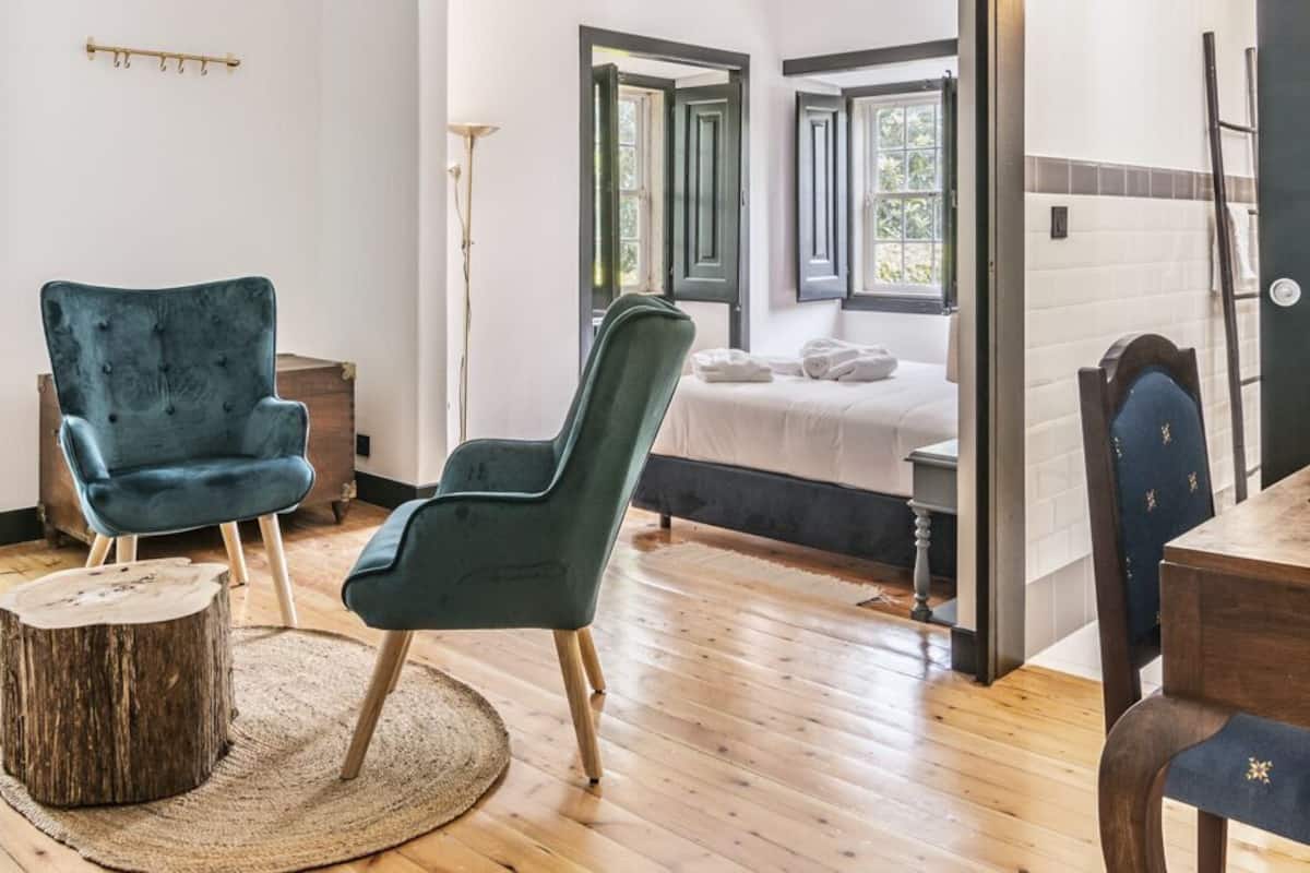 Quarto com uma cama, duas cadeiras, uma mesa de madeira, um banquinho pequeno, uma mesa de madeira redonda e uma janela grande com vista para árvores.