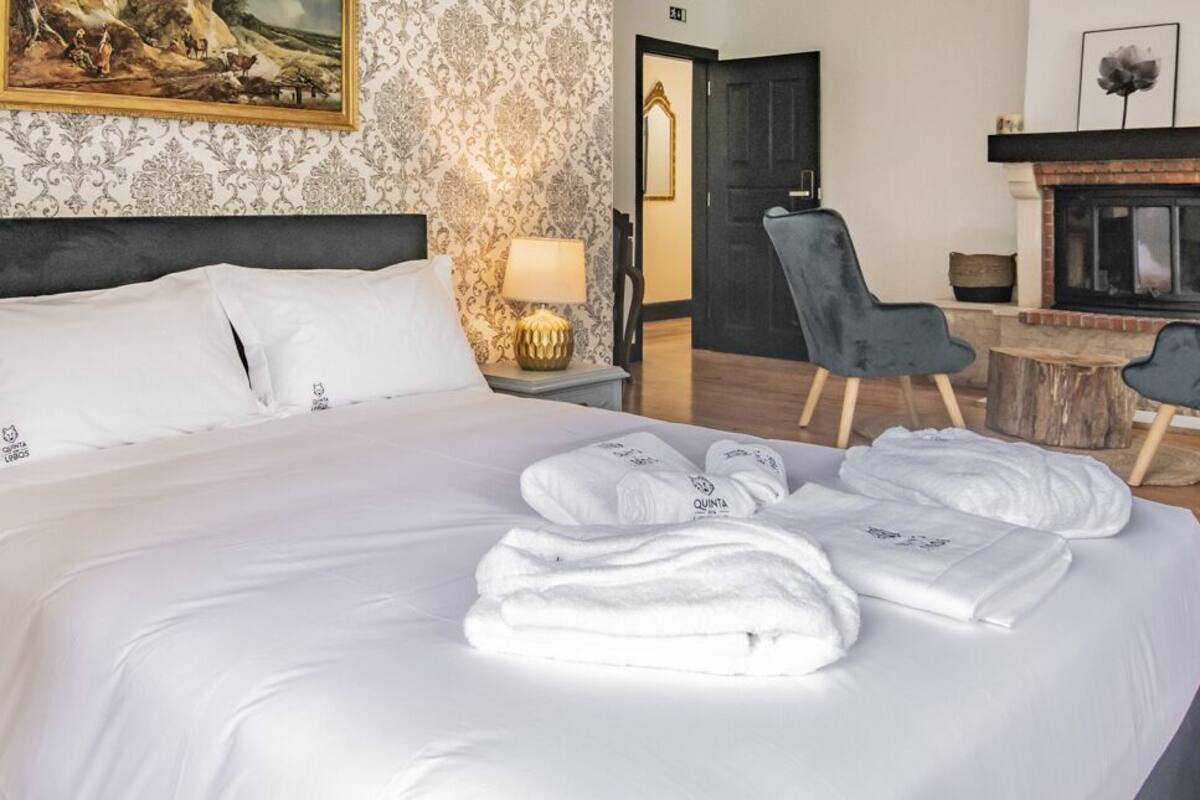 Cama bem arrumada com lençóis e toalhas brancas, cabeceira escura e mesa de cabeceira com abajur.