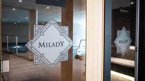 Een glazen deur met erboven een bord waarop 'SAUNA' staat en daarbinnen twee borden met de tekst 'MILADY'.