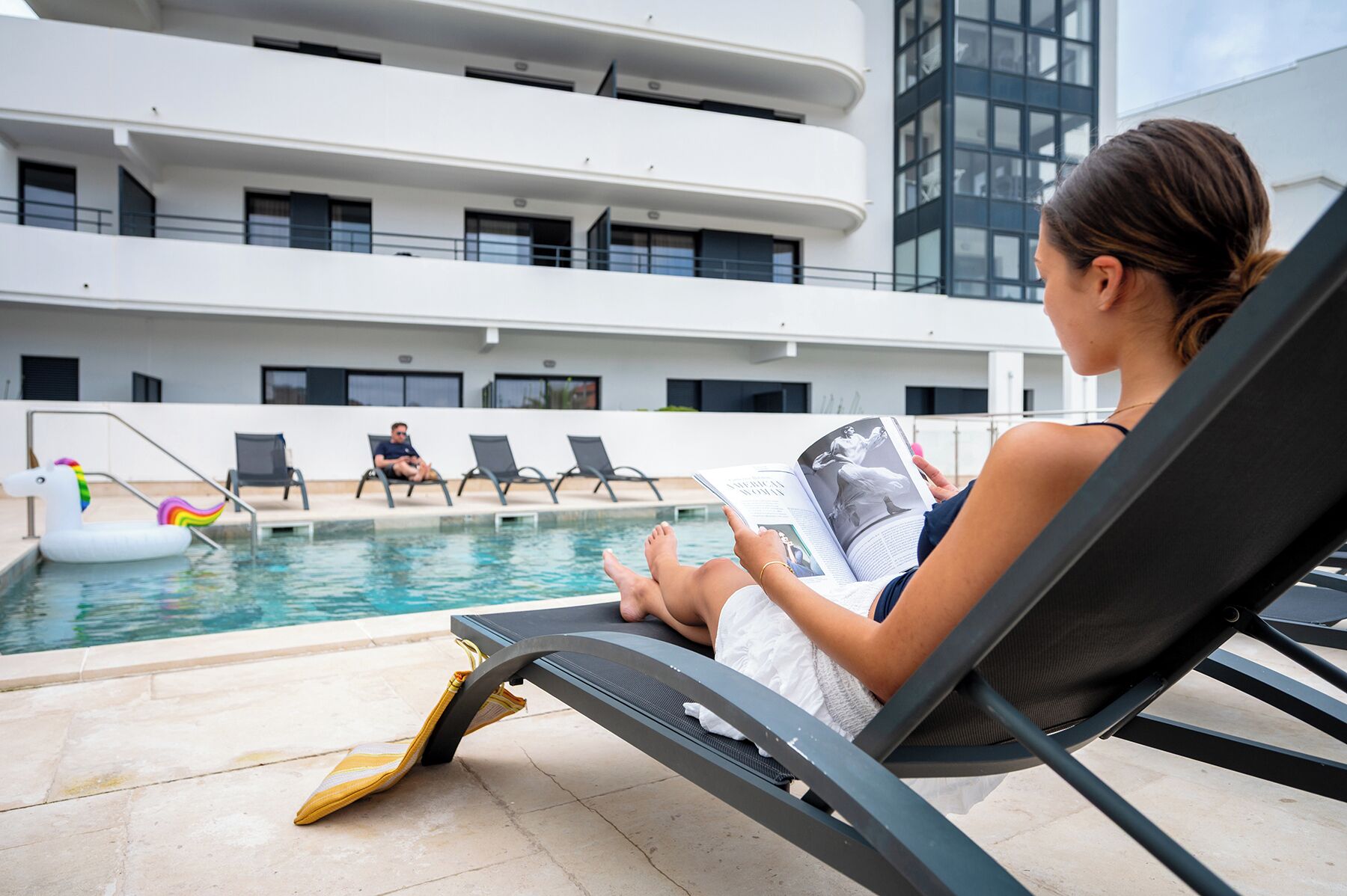 Zona de piscina con sillones de descanso, una piscina y un edificio al fondo.