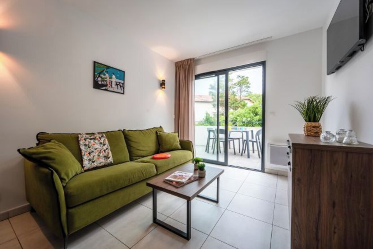 Un canapé vert, un magazine, une petite table, une plante et un meuble dans une pièce dotée d’une porte-fenêtre coulissante donnant sur un espace extérieur.