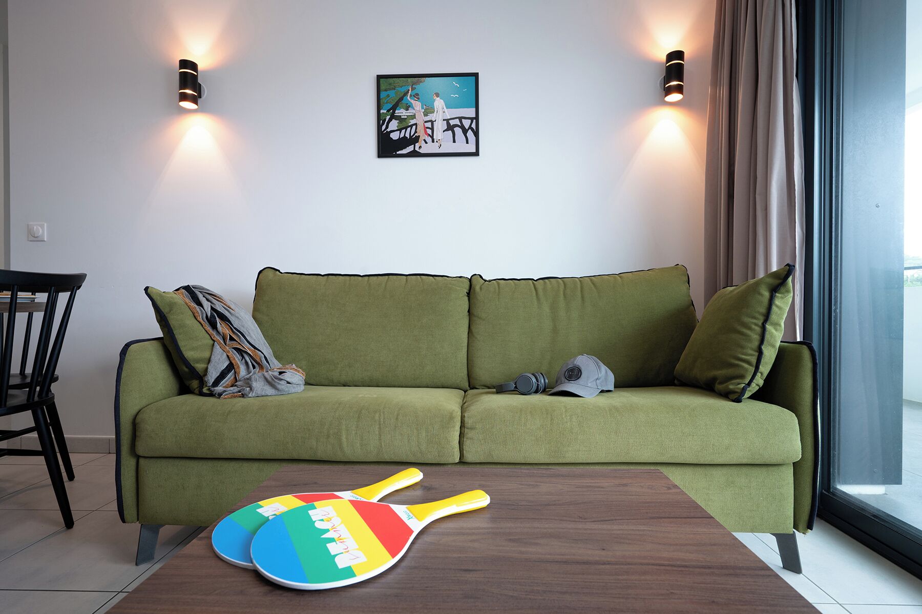 A green sofa with a cushion, a pair of colorful table tennis paddles, and a framed picture on the wall.