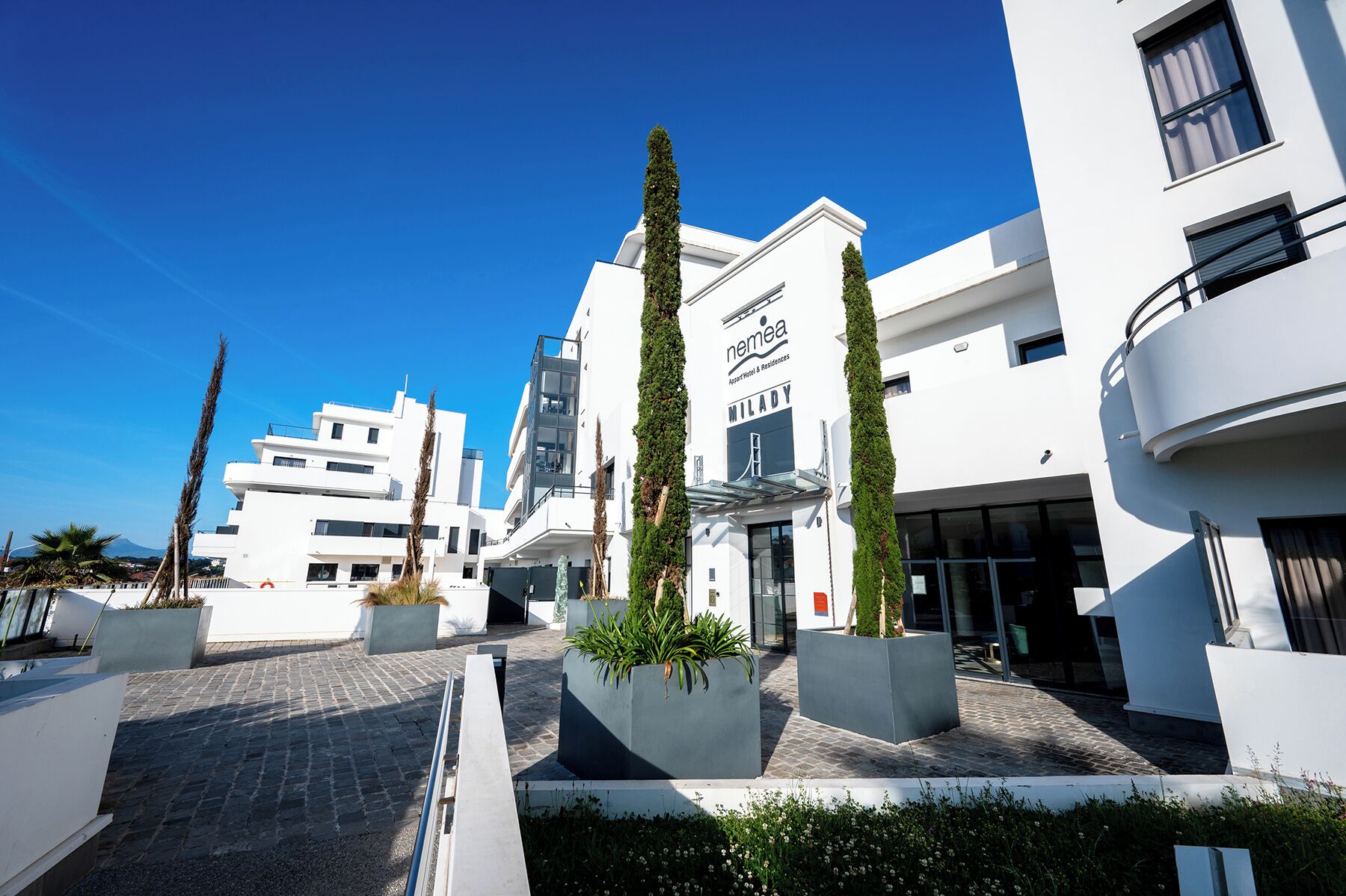 A modern hotel with a clear sky, featuring a prominent entrance with a glass facade, and a sign for 'NEMEA Milady.'