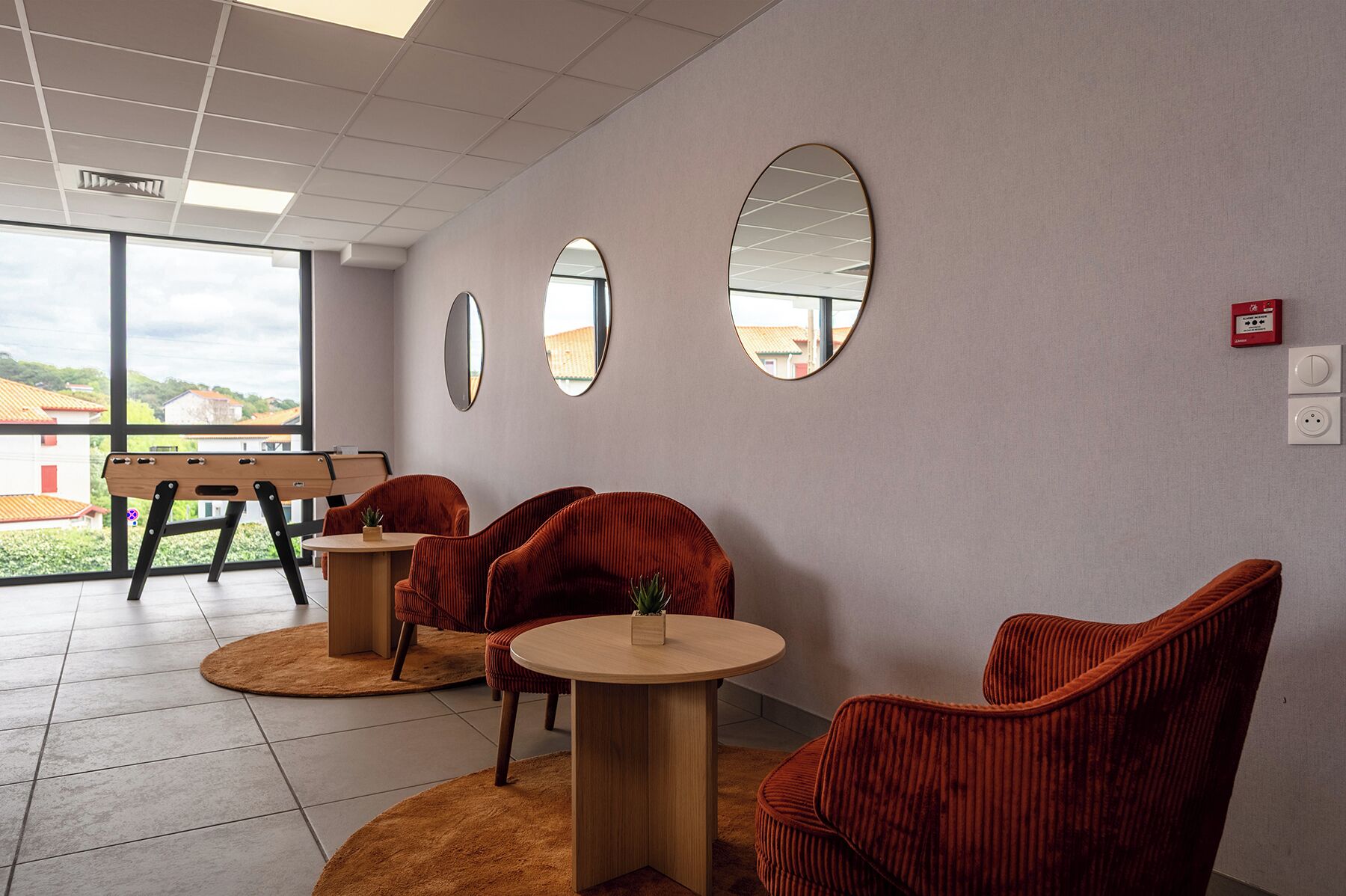 A modern interior with a seating area featuring red upholstered chairs, a wooden table, and a bench. There are three wall-mounted mirrors and a view of the outdoors through large windows.