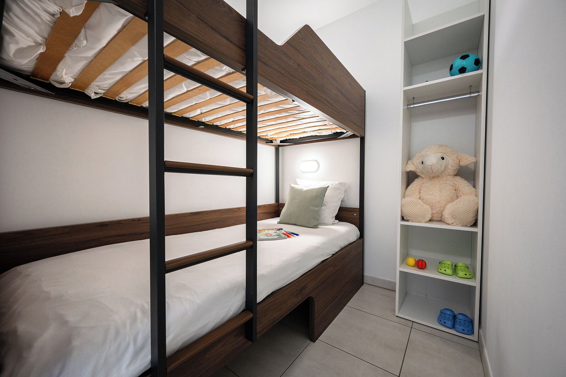 Un letto a castello con struttura in legno e biancheria bianca, uno scaffale con un orsetto e scarpe da bambino, e un piccolo ripiano con un pallone da calcio.