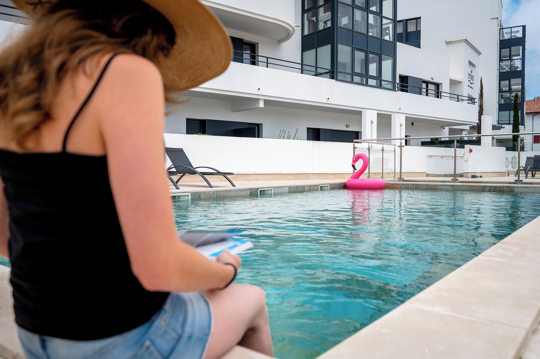 Un área de piscina con un flotador de flamenco rosa, sillones de descanso y un edificio moderno con balcones al fondo.