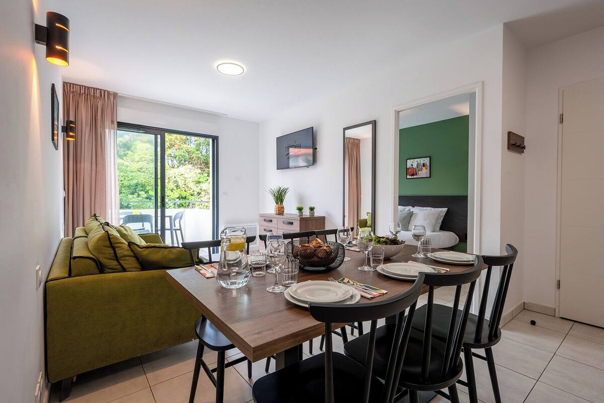 Een eethoek met een houten tafel voor vier personen, omringd door zwarte stoelen. Een bank met groene kussens bevindt zich aan de linkerkant.