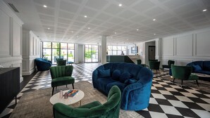 Een ruime lobby met een geruit vloerkleed, zitplaatsen in groen en blauw, een receptiebalie en een modern plafondontwerp.