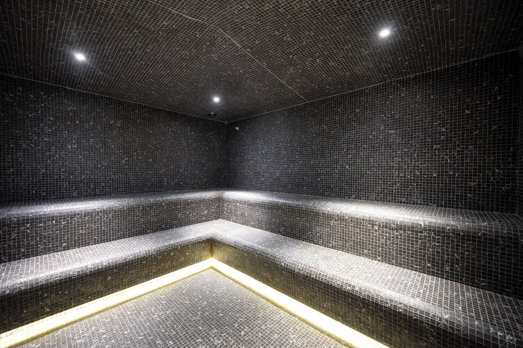 Una stanza moderna, scarsamente illuminata, con una parete rivestita di mosaici strutturati e un'area salotto centrale illuminata da una fascia luminosa.