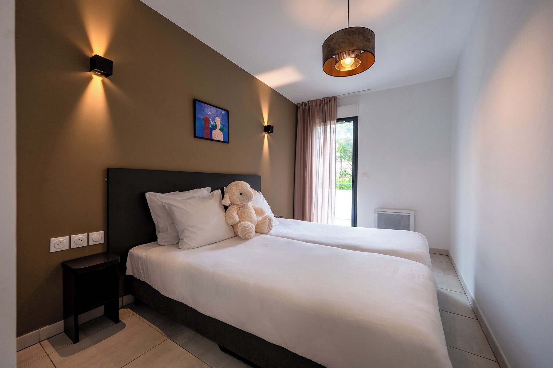 A hotel room with two beds, a nightstand, a wall-mounted light, a ceiling light, a window with curtains, and a painting on the wall.
