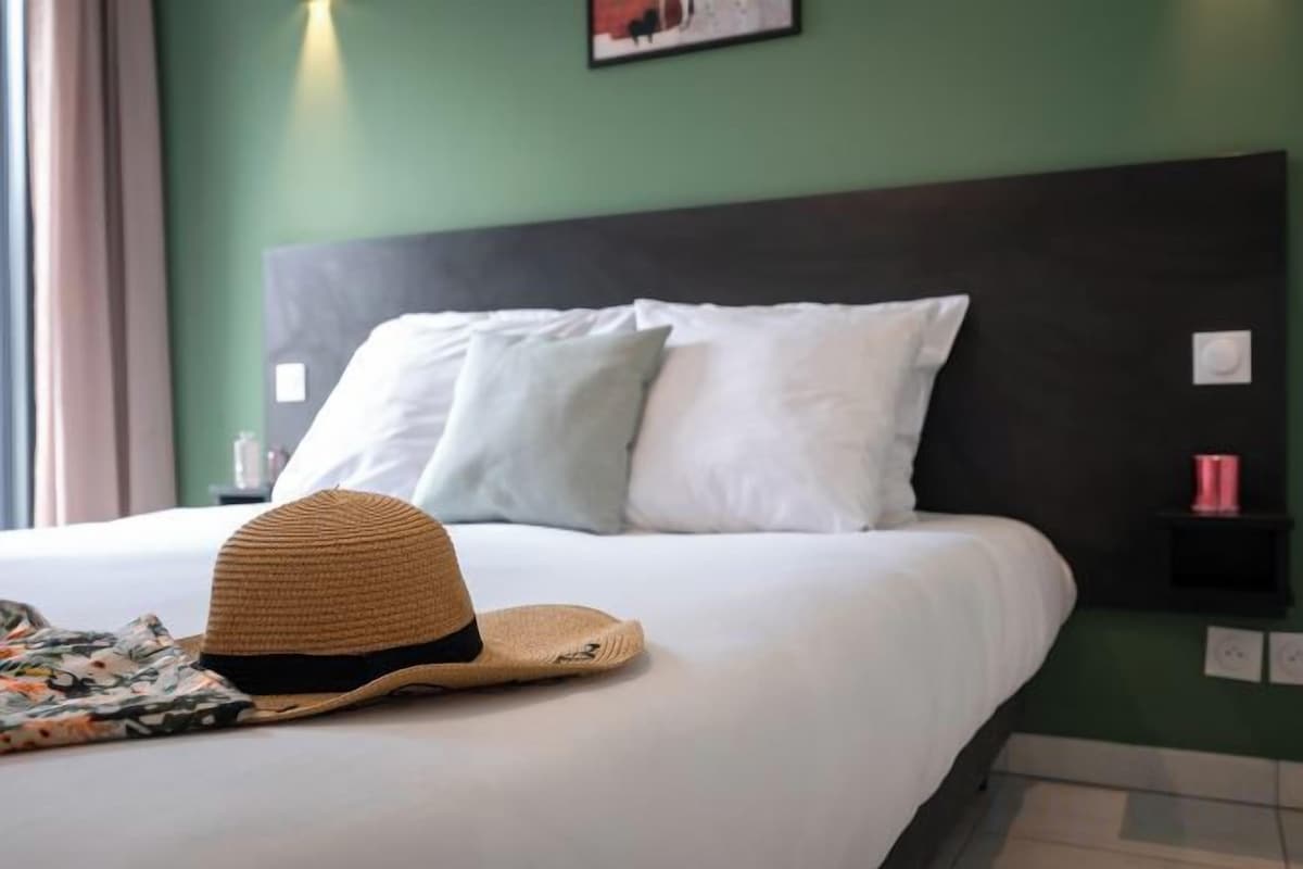 Una cama con ropa blanca, un sombrero de paja y una mesita de noche con una taza roja.