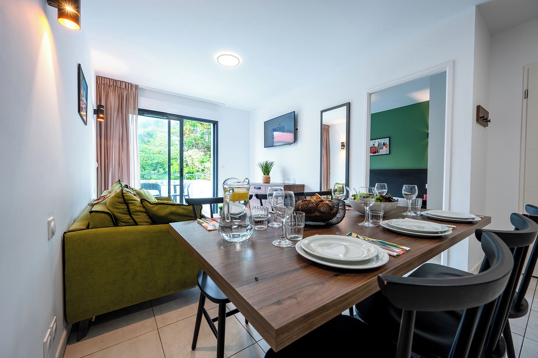 Un comedor con una mesa de madera dispuesta para cuatro personas, un sofá verde y un amplio ventanal con cortinas.