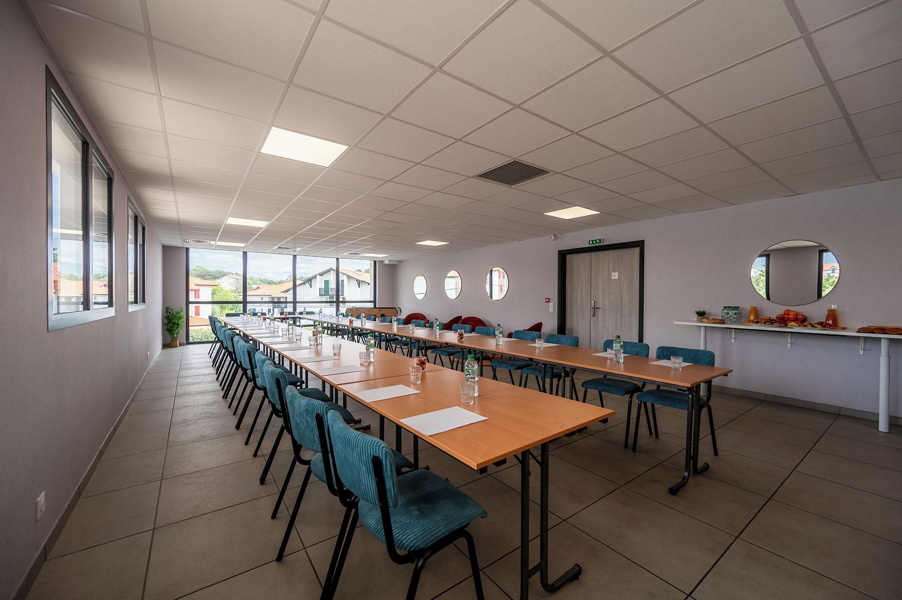 Une salle de conférence équipée de longues tables, de chaises et d’une grande fenêtre offrant une vue sur les bâtiments environnants.