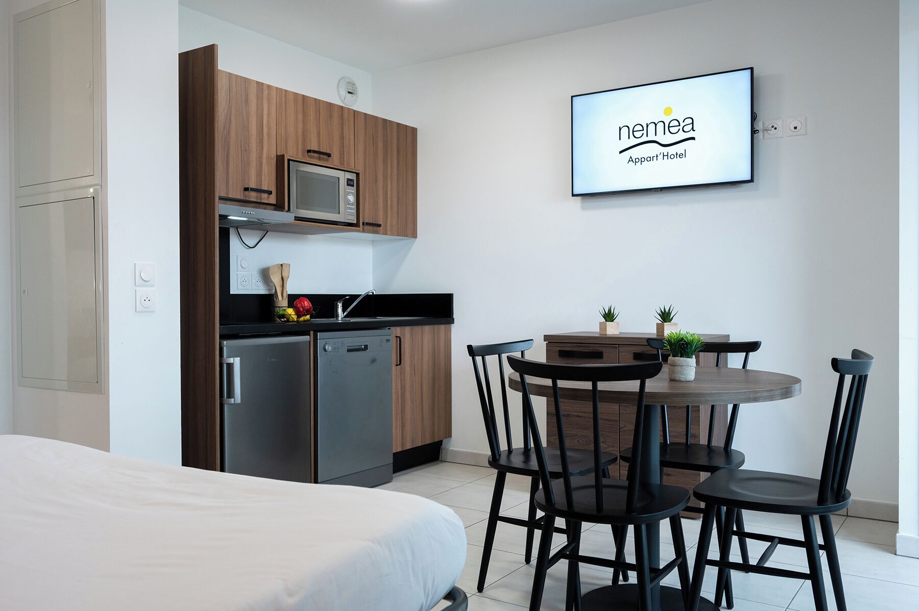 A compact kitchenette with a microwave, a small refrigerator, and a sink. There is a dining area with a round table and four chairs. A flat-screen TV is mounted on the wall displaying the logo of 'nemea Apart'Hotel'.