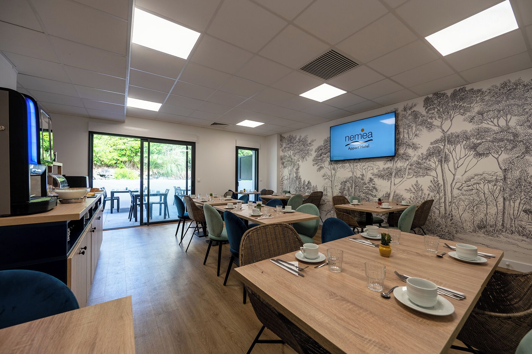 A conference room with tables set for a meeting, a coffee machine, and a television screen displaying the Nemea Appart'Hotel logo.
