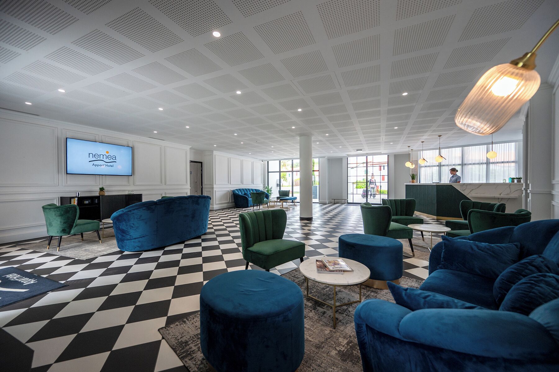 Een ruime lobby met een zwart-wit geruit vloerkleed, blauwe banken, groene stoelen en een receptiebalie.