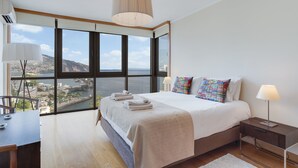 A modern hotel room with a large window offering a city view, a neatly made bed, a desk, and a lamp. - Funchal View Apartment (Funchal)