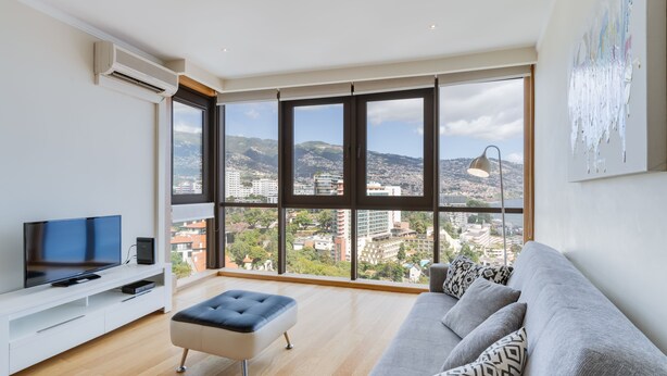 A modern living room with a large window, a flat-screen TV, a sofa, and a small ottoman. - Funchal View Apartment (Funchal)
