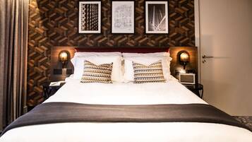 Una cama bien hecha con almohadillas estampadas, cabecera con luces fijadas a la pared y cuadros en la pared.