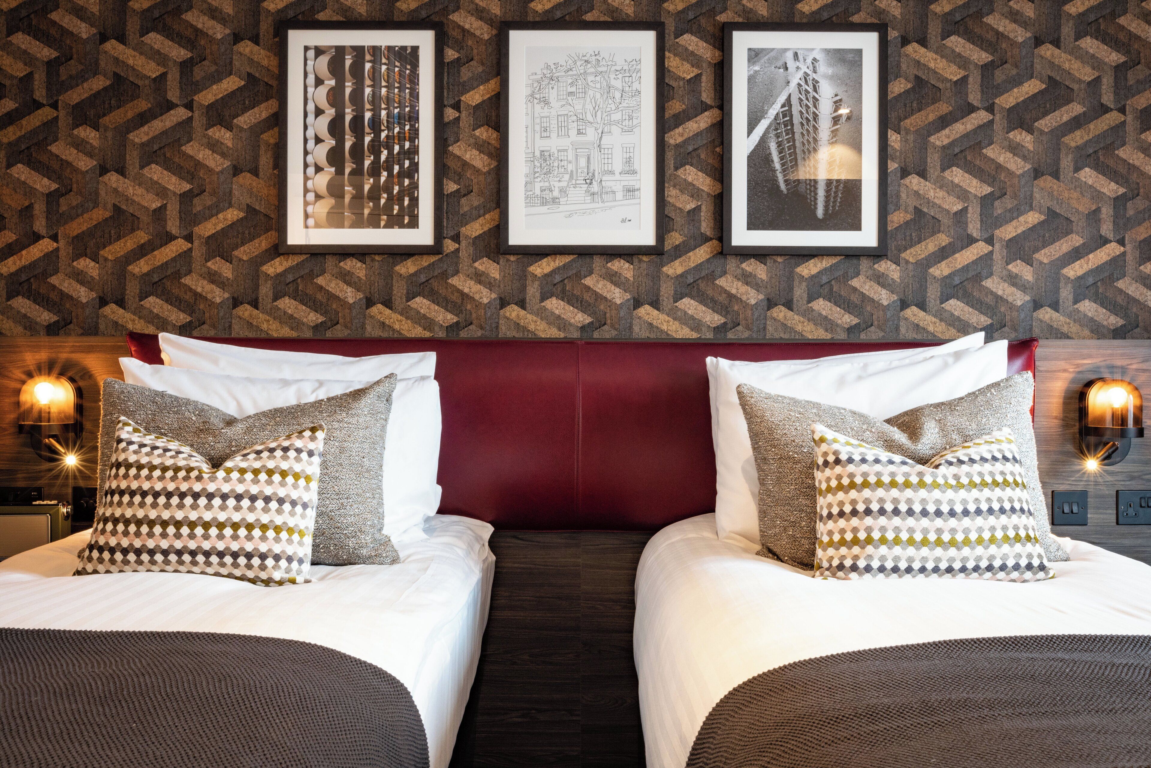 A hotel room with two beds, a red headboard, patterned pillows, and framed artwork on the wall.
