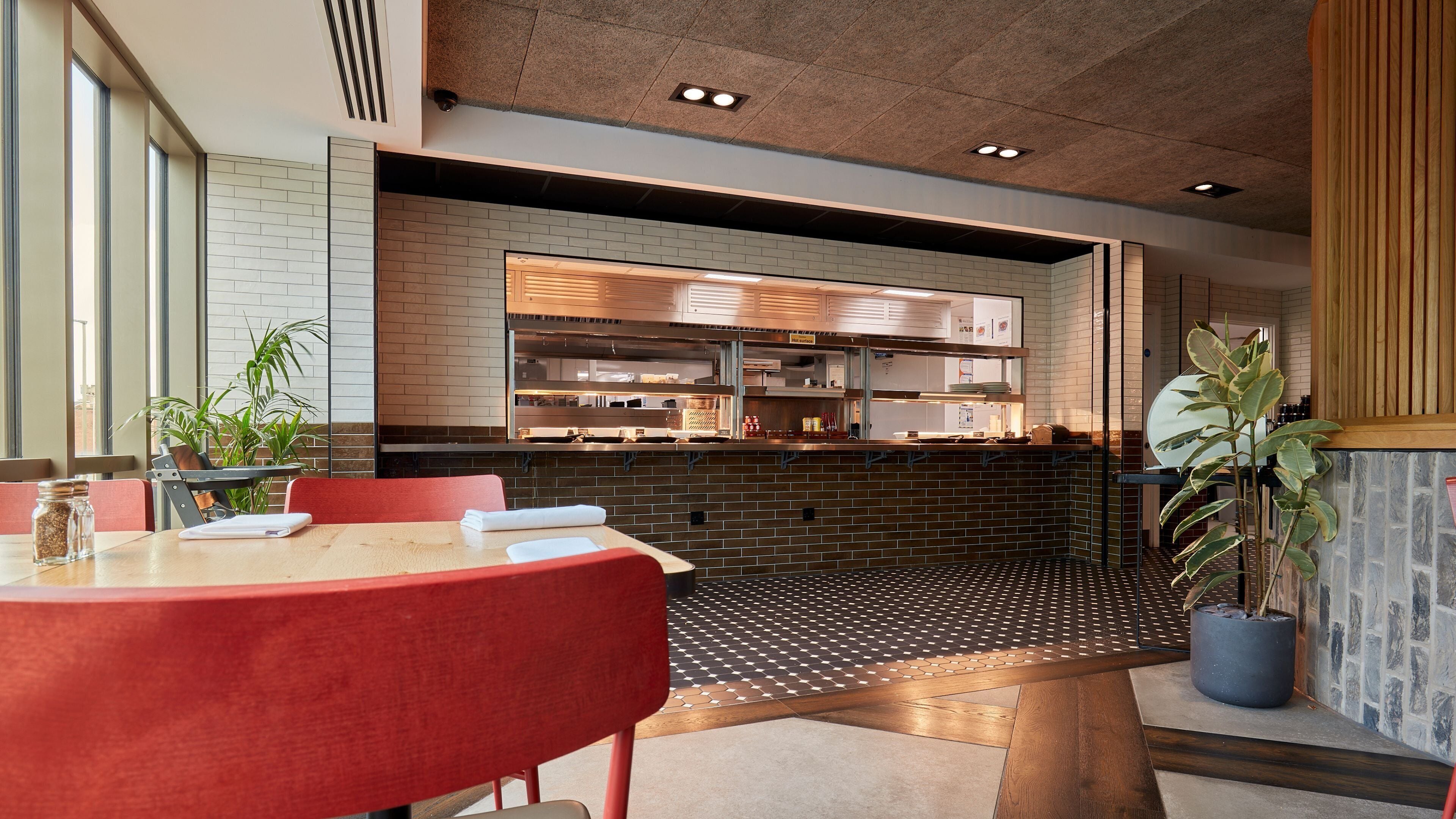 A restaurant with a brick wall, a kitchen area, and a seating area with red chairs and a wooden table.