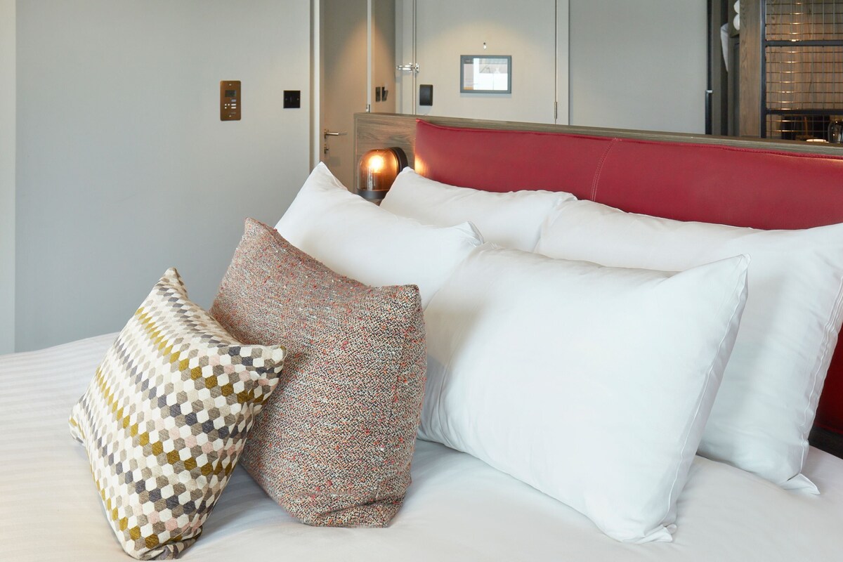 A neatly made bed with white and patterned pillows, a red headboard, and a wall-mounted control panel.
