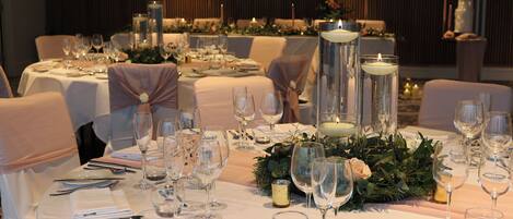 Un salón de banquetes con mesas redondas dispuestas para un evento formal, que incluye cristalería, cubiertos y velas.