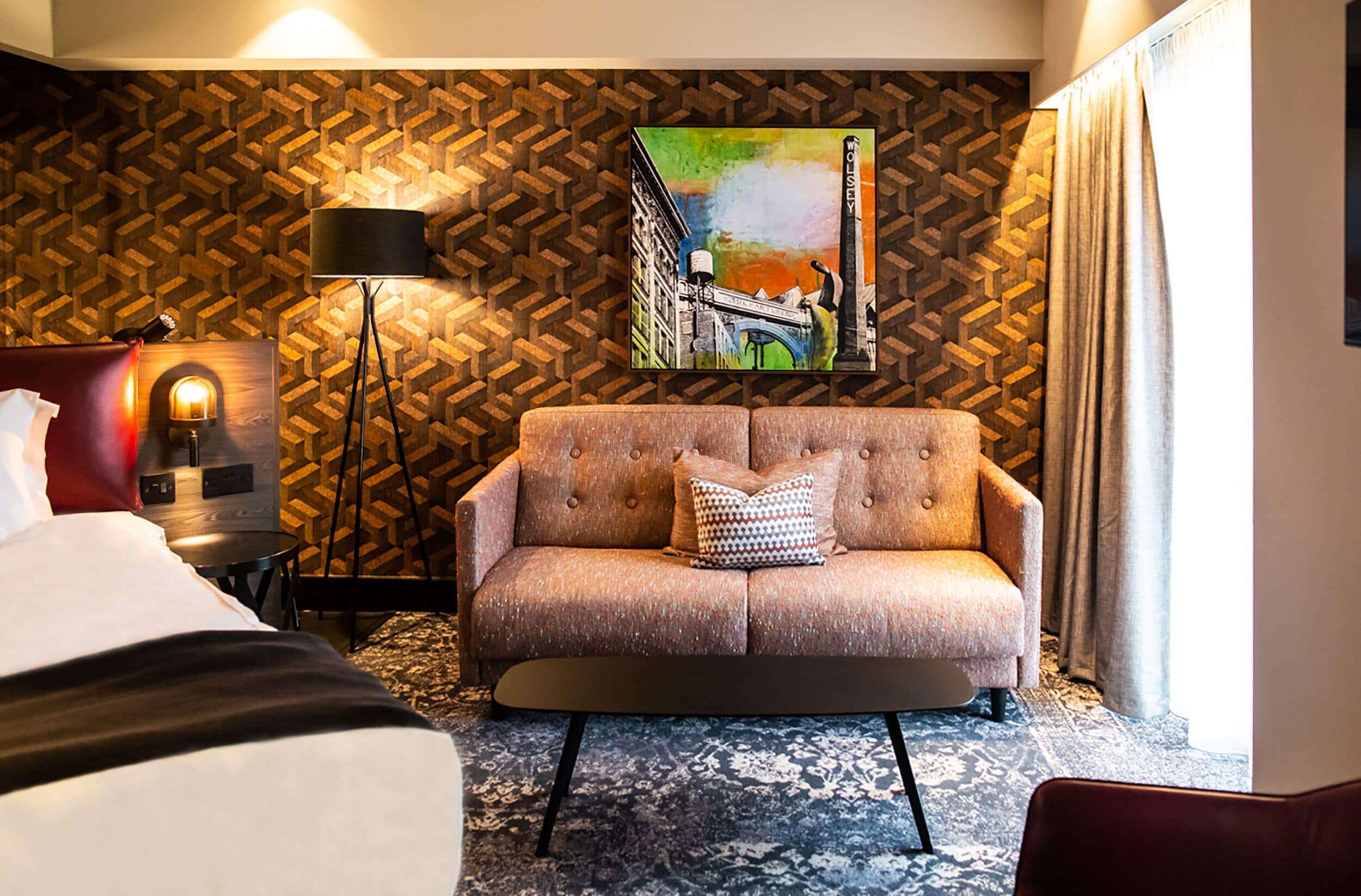 A hotel room with a patterned herringbone wall, a large painting of a bridge, a tufted sofa, a bedside table with a lamp, and a bed with a red headboard.