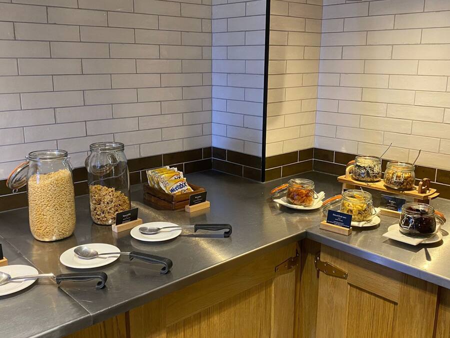 Una barra de buffet con varios alimentos en frascos y cuencos, identificados con nombres como 'Granola' y 'Special K'.