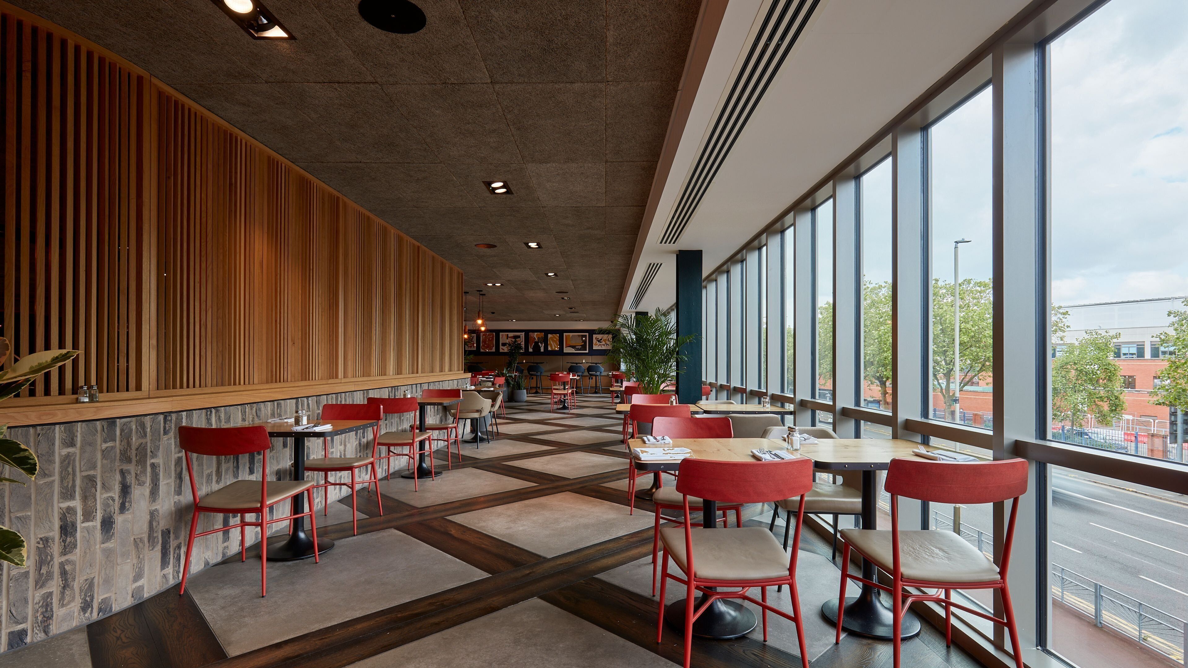A modern restaurant interior with a mix of red and beige chairs, a stone wall, and large windows offering a view of the city.