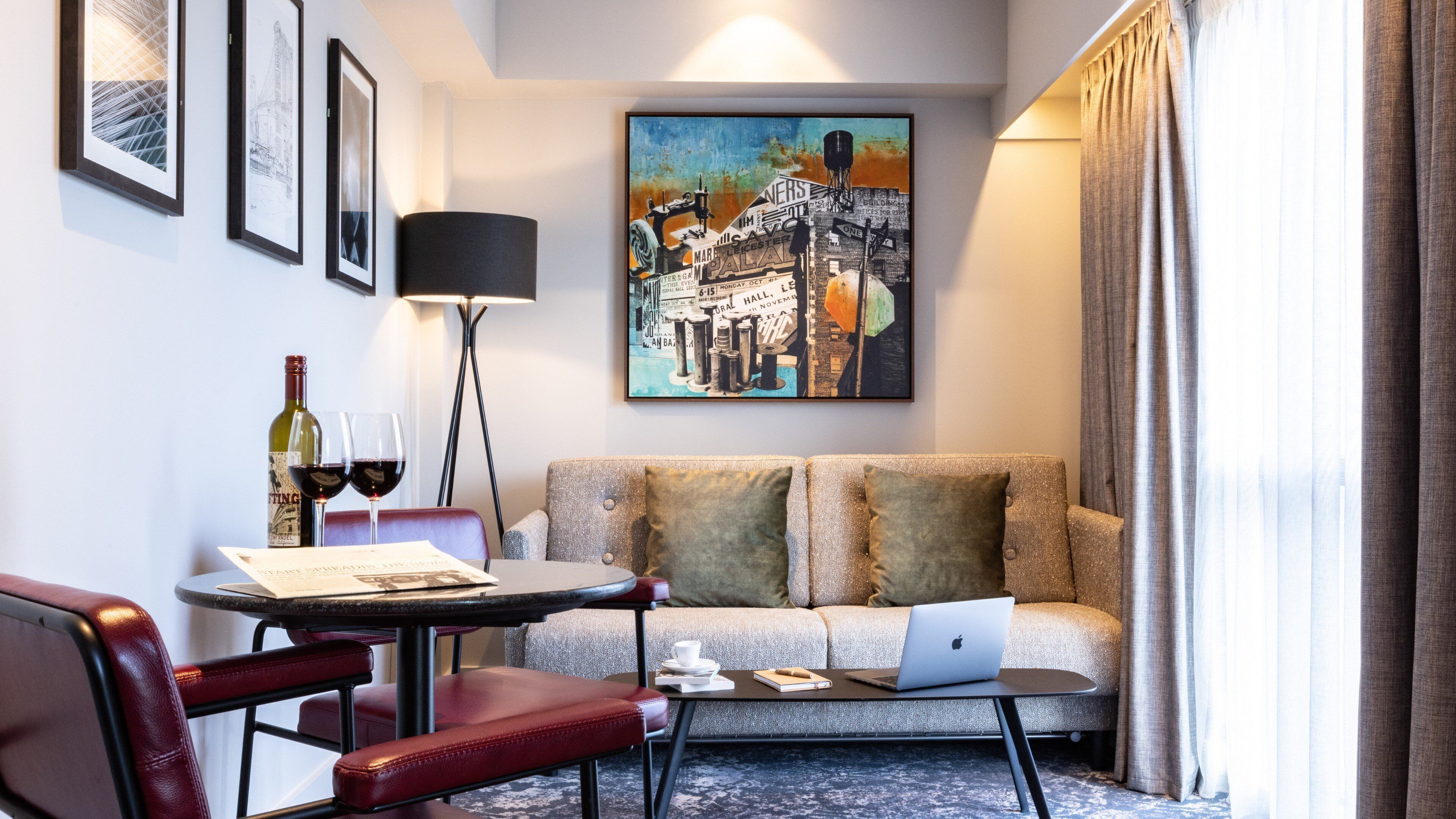 Une chambre d’hôtel avec un canapé, une table à manger avec des chaises, un ordinateur portable, une bouteille de vin et des œuvres d’art encadrées accrochées au mur.