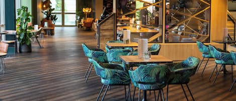 Una cafetería moderna con mesas y sillas de madera, una escalera con diseño geométrico y plantas en macetas.