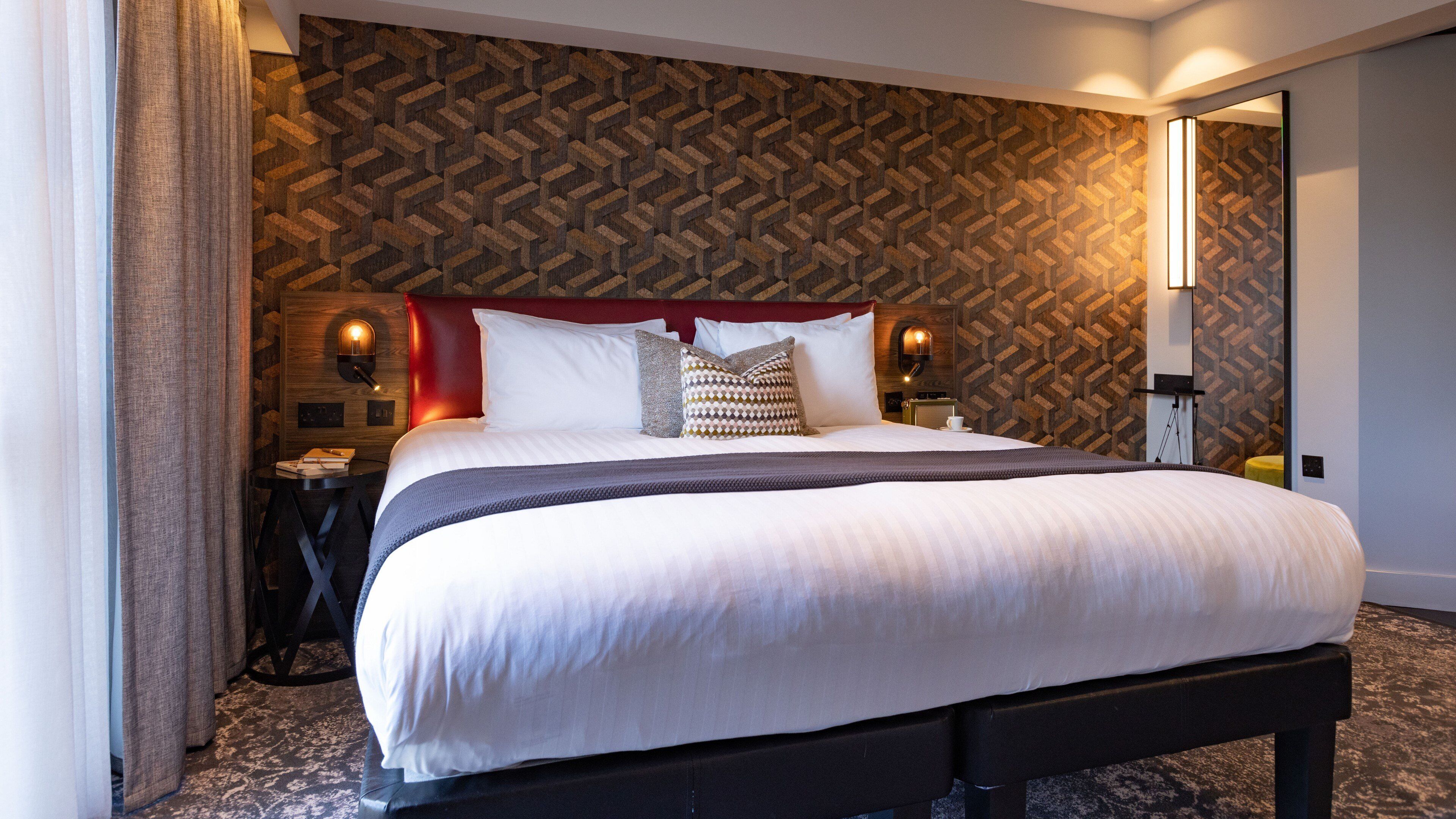 A neatly made bed with a headboard, a bedside table with a lamp, and a patterned wall.