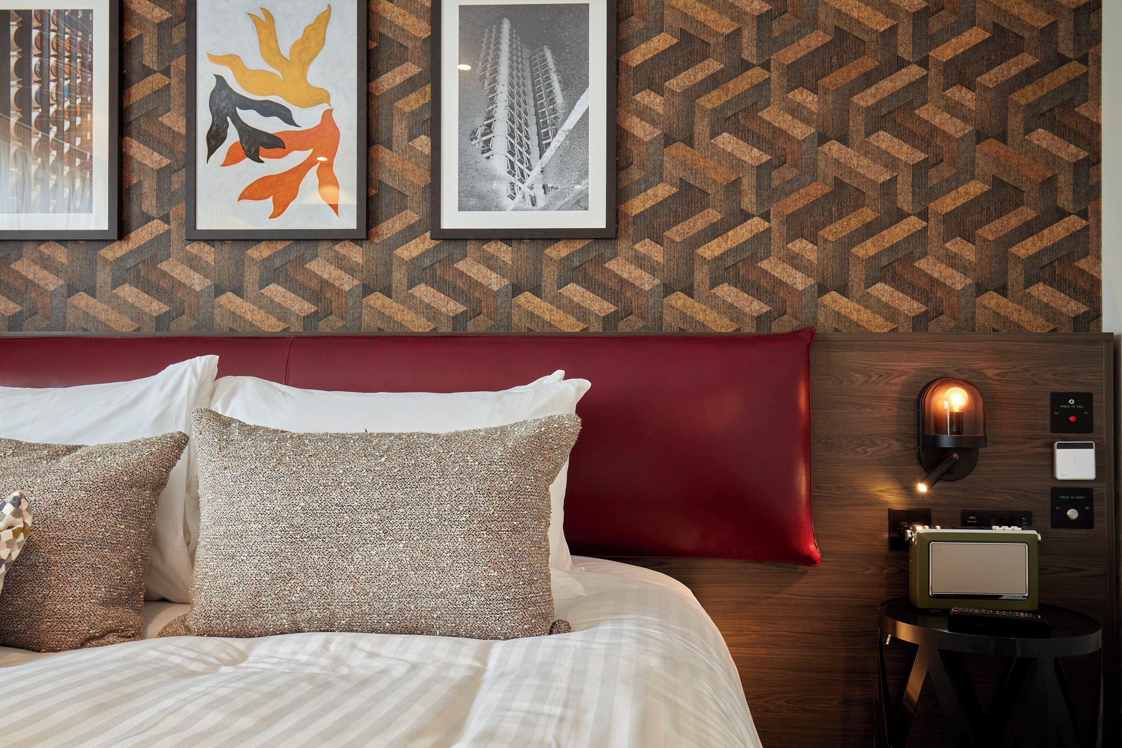 Una cama bien tendida con sábanas blancas y almohadones decorativos, cabecera roja y papel tapiz con diseño geométrico.