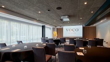Ett konferensrum med flera bord, stolar, en projektorduk som visar texten ”VOCO AN IHG HOTEL” och stora fönster med tunna, genomskinliga gardiner.