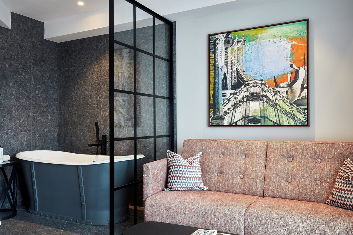 A modern bathroom with a freestanding bathtub, a sofa with patterned cushions, and a large abstract painting on the wall.
