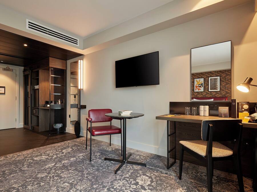A hotel room with a desk, two chairs, a flat-screen TV, a mirror, and a small table with a lamp.