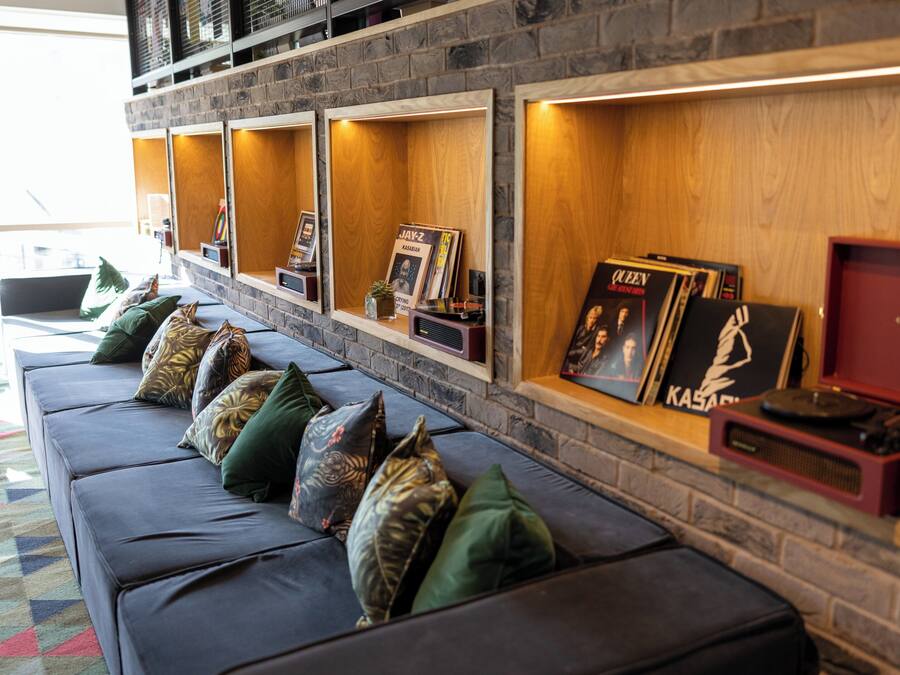 Ein stilvoller Lounge-Bereich mit einer Ledersofa, grünen und gemusterten Kissen sowie einem an der Wand befestigten Regal, das gerahmte Schallplatten und einen Vintage-Plattenspieler zeigt.