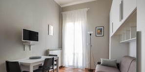 Ein kleiner, hell erleuchteter Raum mit einem weißen Schreibtisch, einem Stuhl, einem Fernseher, einem Heizkörper, einer Stehlampe und einem Sofa.