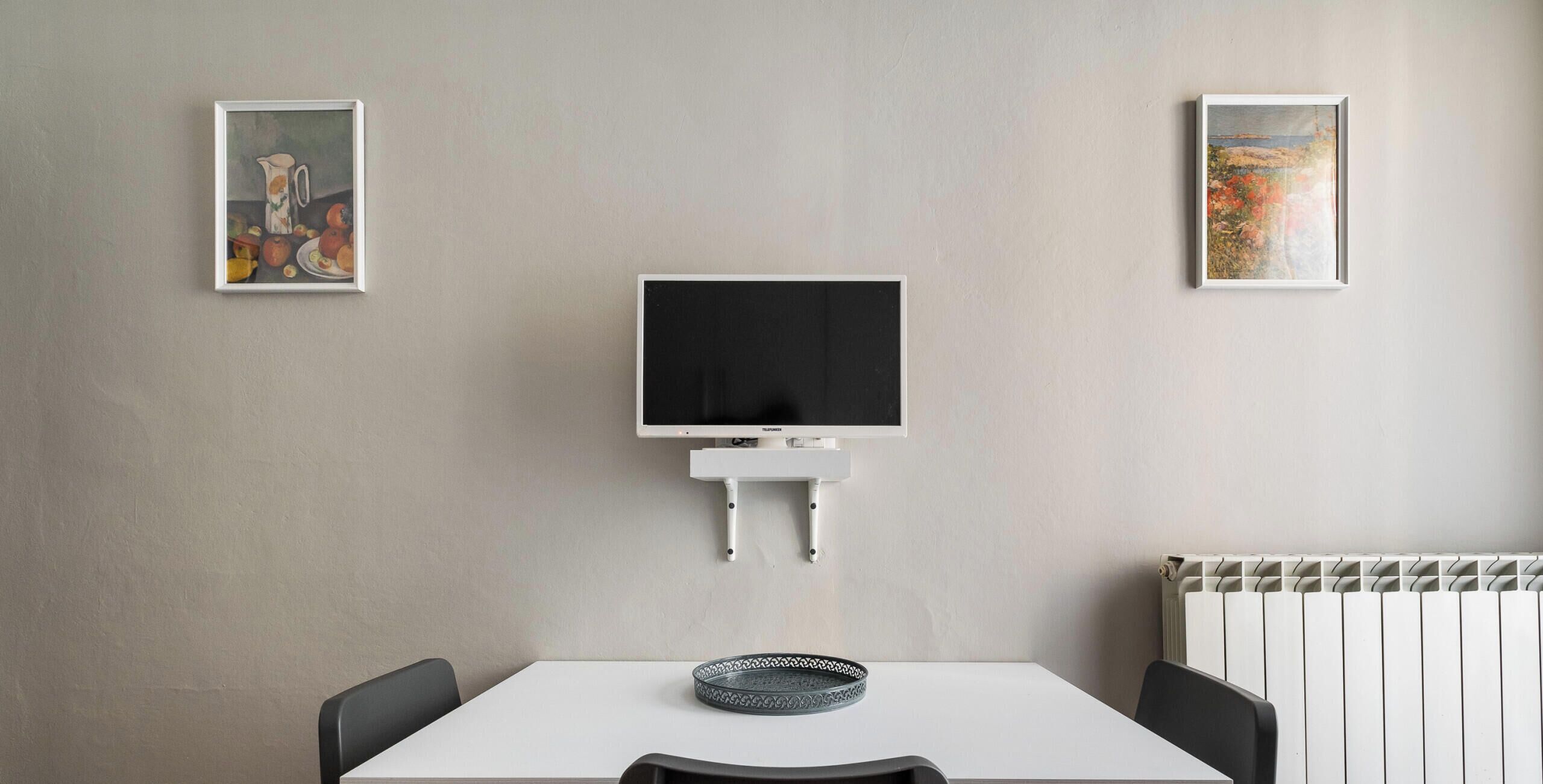 Et moderne, minimalistisk værelse med et hvidt bord, to sorte stole, et fladskærms-tv monteret på væggen og to indrammede kunstværker på væggene.