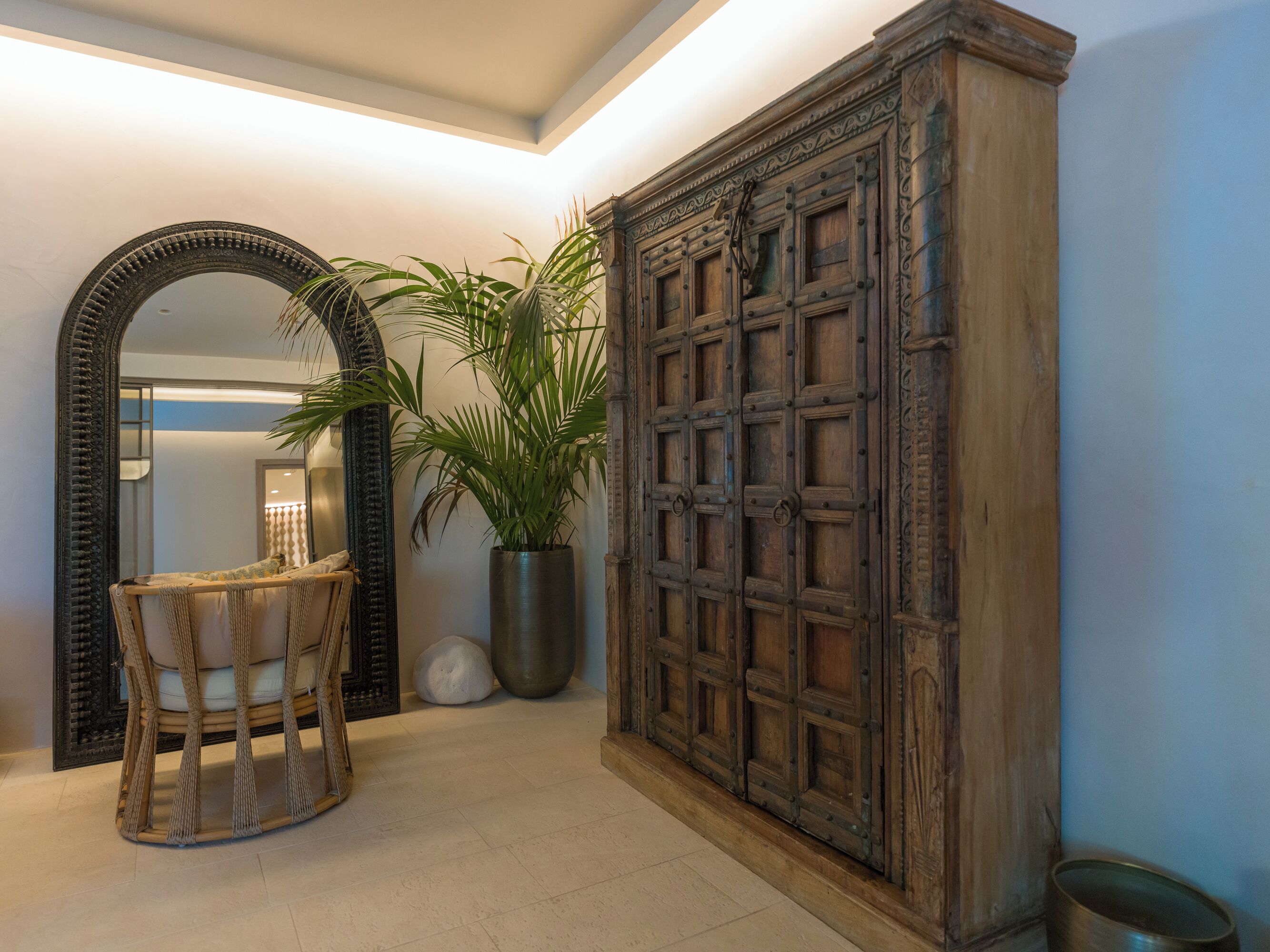 A hallway with a large wooden cabinet, a mirror, a potted plant, and a wicker chair.